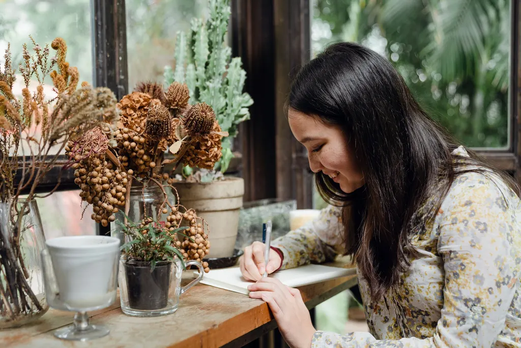 A smiling woman writes in a notebook at a sunny table by the window.