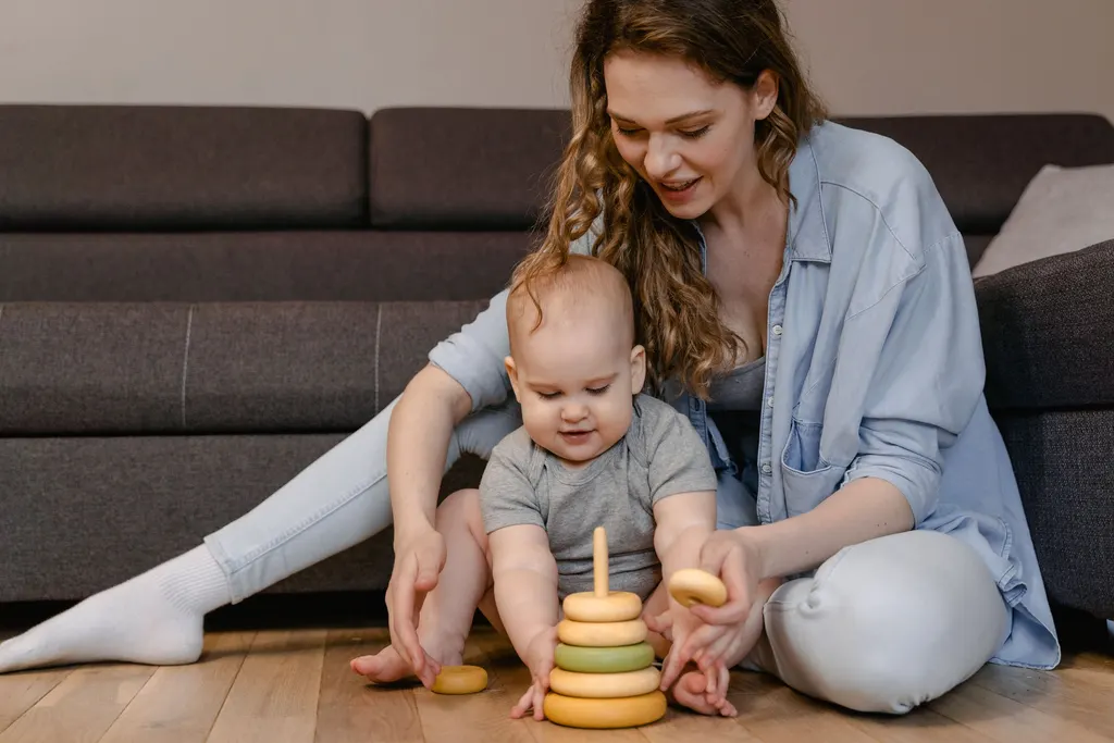 A woman sits on the floor playing with a baby and a wooden stacking toy.