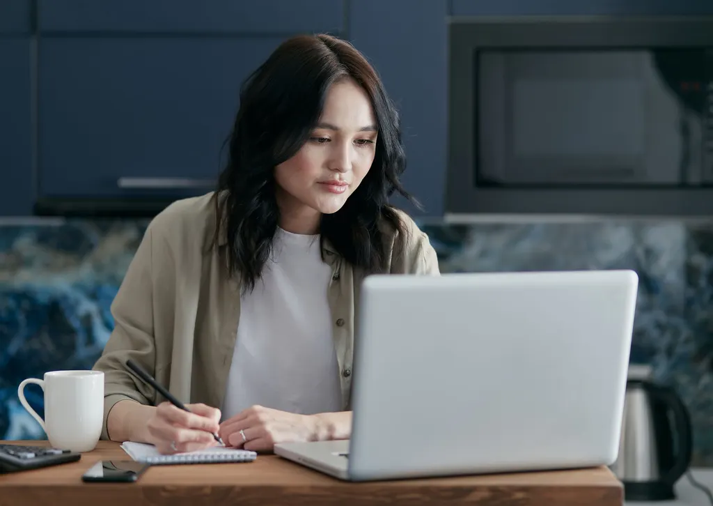 A woman takes notes beside her laptop at the kitchen counter.