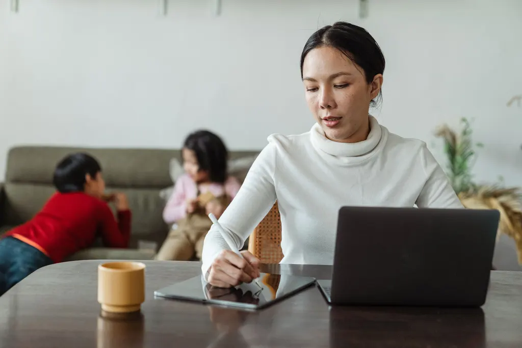 A mother works at her laptop at the dining table while two children play on the sofa behind her.