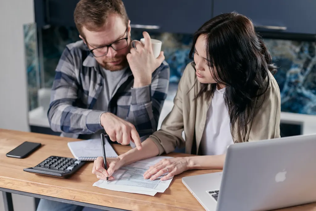 A couple goes over household paperwork with a calculator and a laptop at the kitchen table.