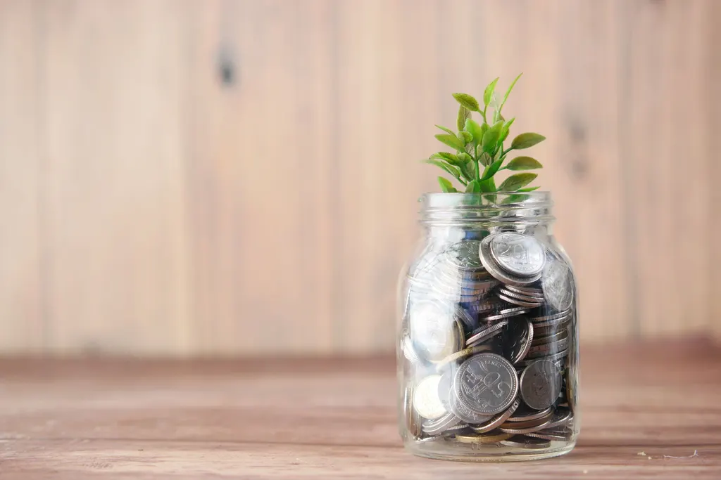 A glass jar of coins with a green sprout growing from it, on a wooden table.