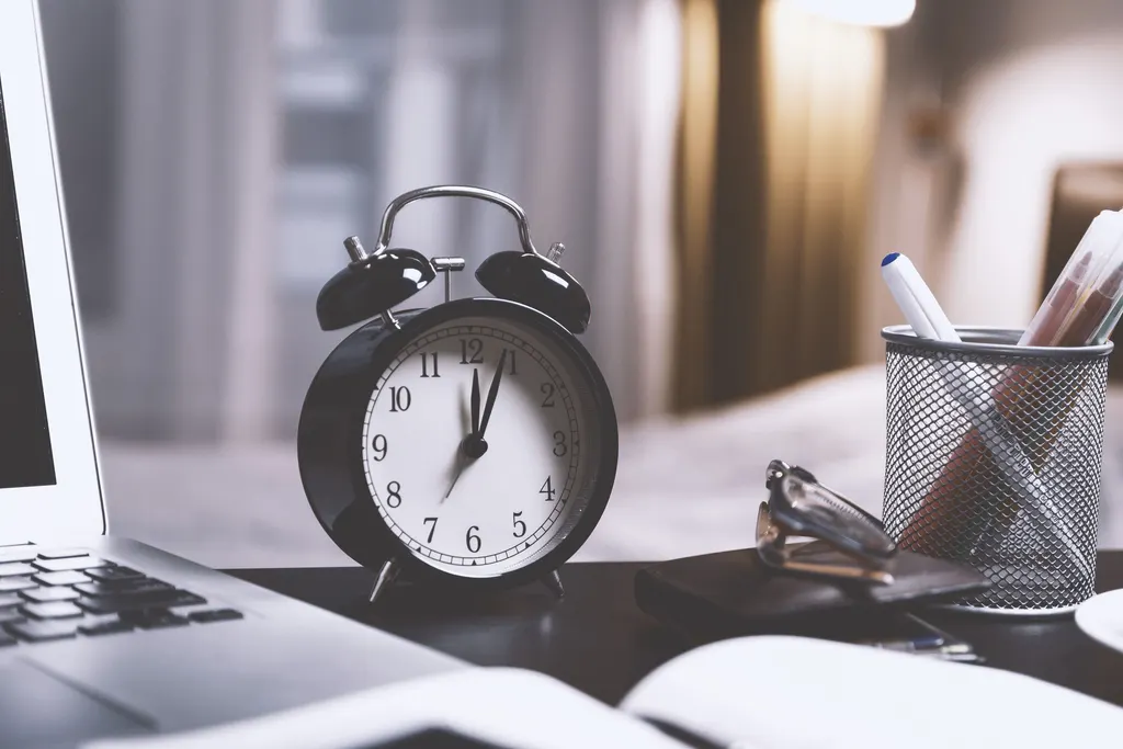 An alarm clock on a desk beside a laptop and a cup of pens.