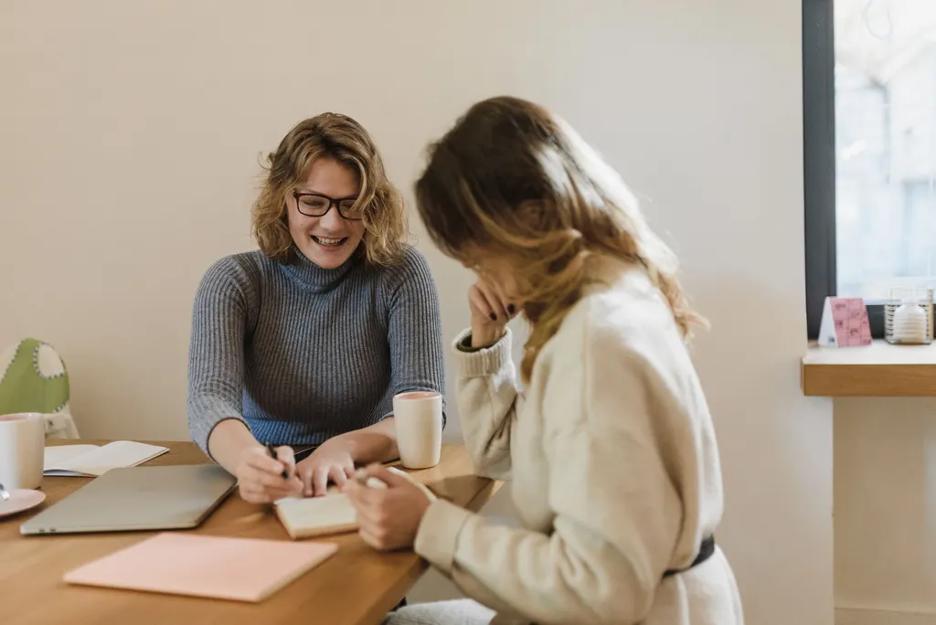 Two women laughing as they talk over notebooks at a wooden table.