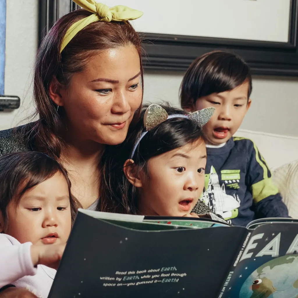 A woman reads a picture book with two young children on the sofa.